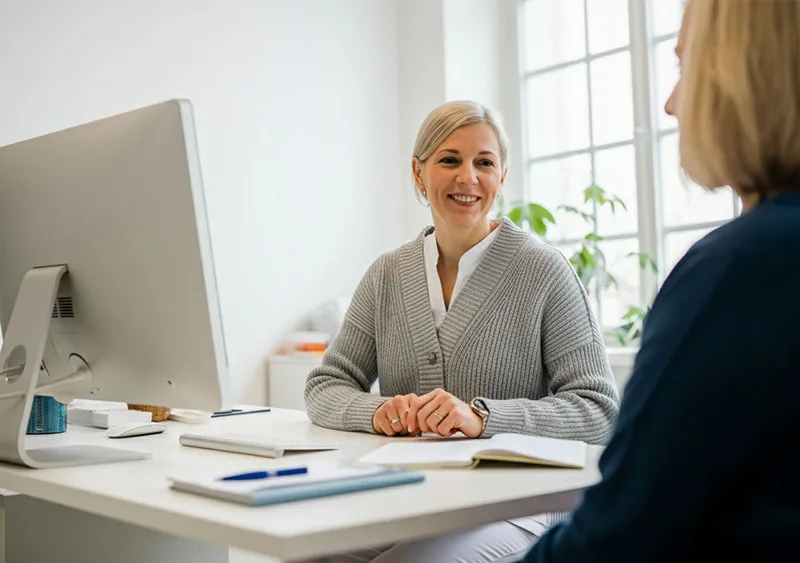 Eine freundliche Mitarbeiterin im Büro von SANICUR Dresden GmbH sitzt lächelnd an einem weißen Schreibtisch und führt ein Beratungsgespräch mit einer Person, die von hinten zu sehen ist. Die Mitarbeiterin trägt eine graue Strickjacke über einer weißen Bluse. Vor ihr steht ein Computerbildschirm, und auf dem Tisch liegen Notizblöcke und Stifte. Im Hintergrund ist ein großes Fenster zu sehen, das viel Licht in den Raum lässt, und eine grüne Pflanze. Die Szene wirkt hell und einladend und vermittelt eine professionelle, offene Atmosphäre für Gespräche und Beratung.